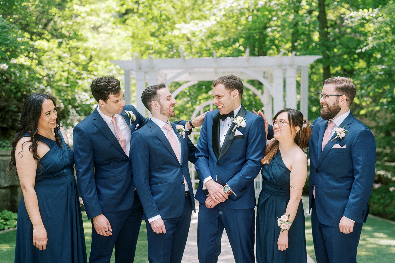 Rawle smiles with bridesmaids and groomsmen in navy attire beneath a white garden pergola.
