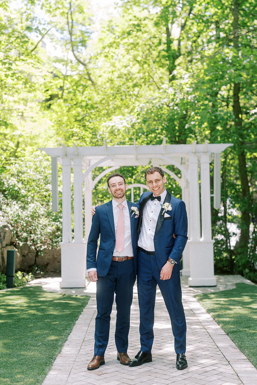 Two men in blue suits pose arm in arm beneath a white garden arbor at Ancaster Mill.