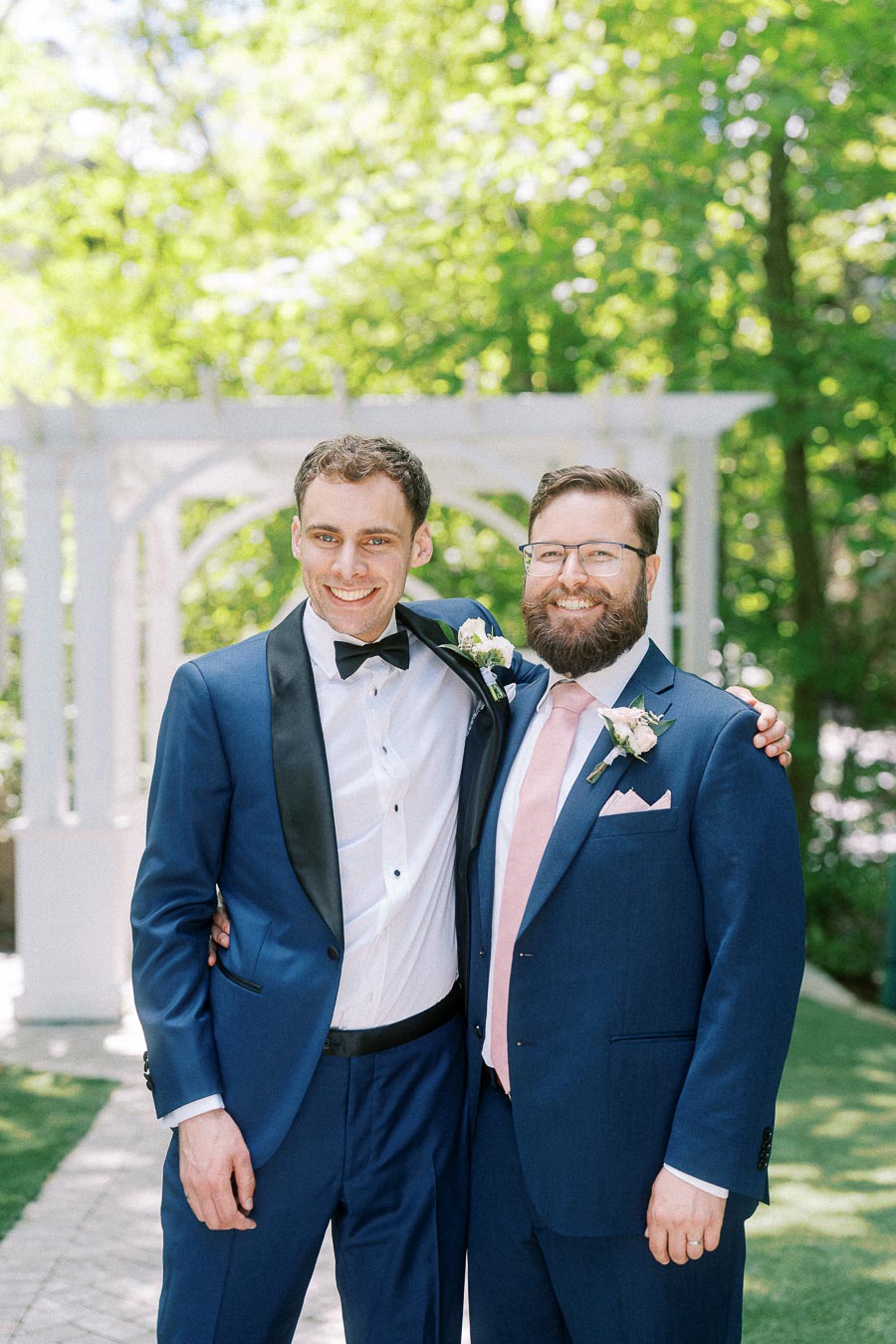 Two men in blue suits smile with arms around each other near a white garden arbor.