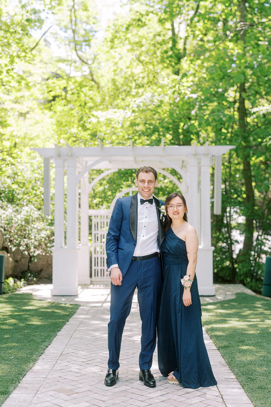Wedding guest couple in navy formal attire standing on a garden path before a white arbor.