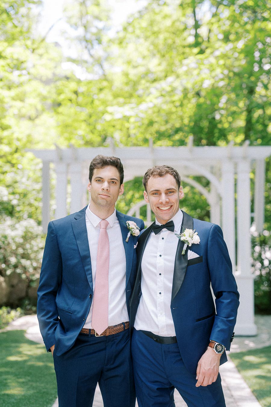 Two men in blue suits pose together outdoors beneath a white pergola surrounded by green trees.