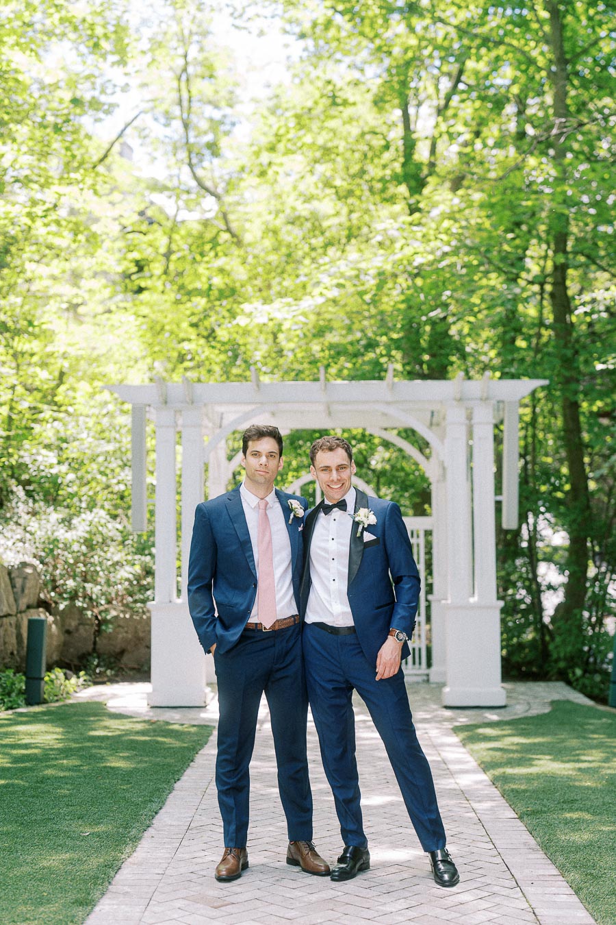 Two men in blue suits stand together on a garden path before a white arbor at Ancaster Mill.