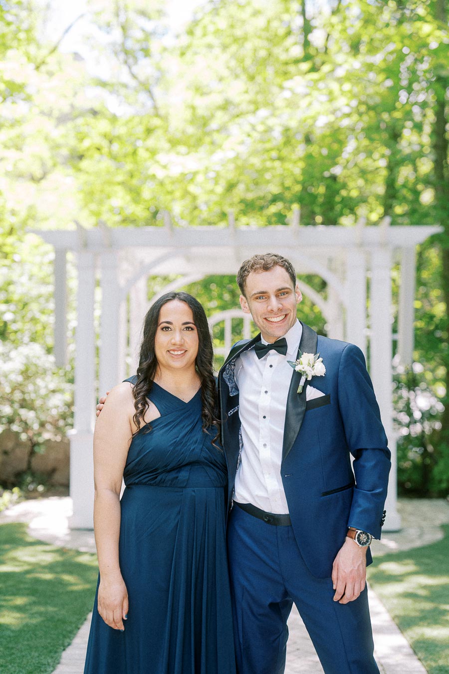 Wedding attendant in navy dress stands with groomsman in blue tuxedo before a white garden pergola.