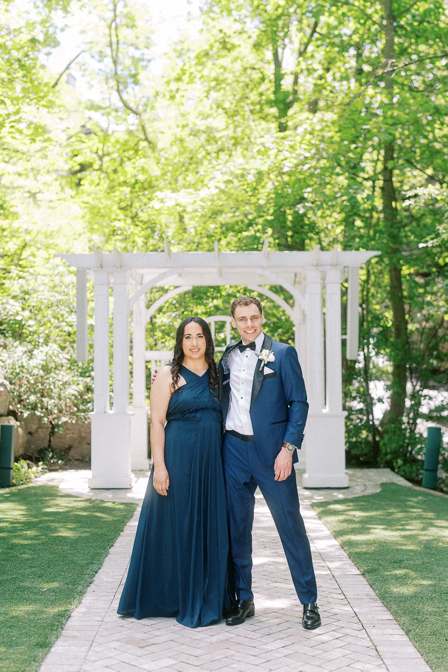 Wedding party pair in navy formalwear standing on a garden walkway beneath a white pergola.