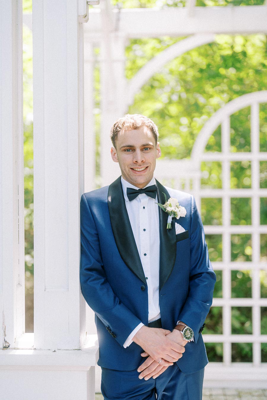 Groom Rawle in a blue tuxedo and black bow tie standing by white garden arches in bright greenery.