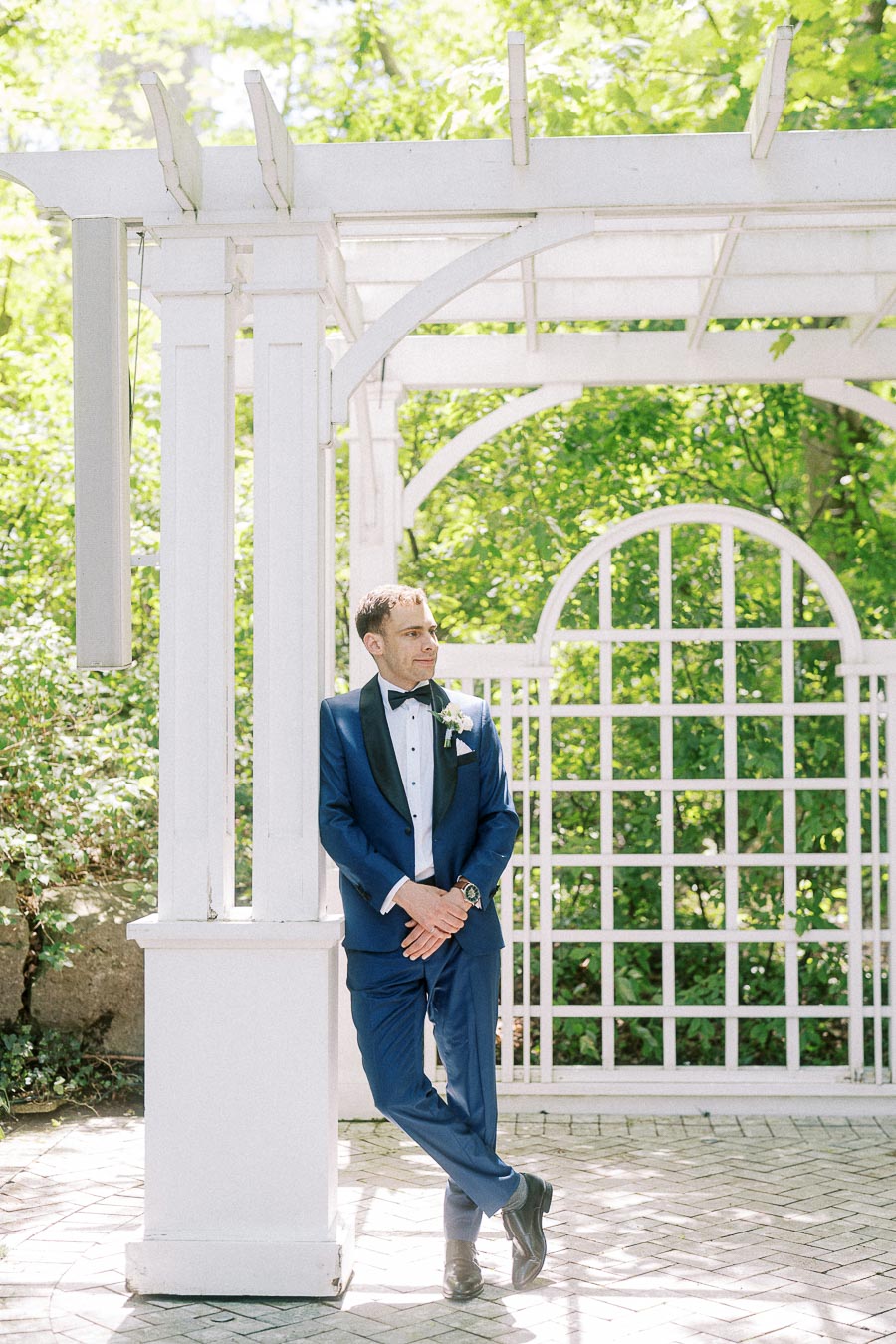 Groom in a blue tuxedo leaning against a white pergola in a sunny garden setting