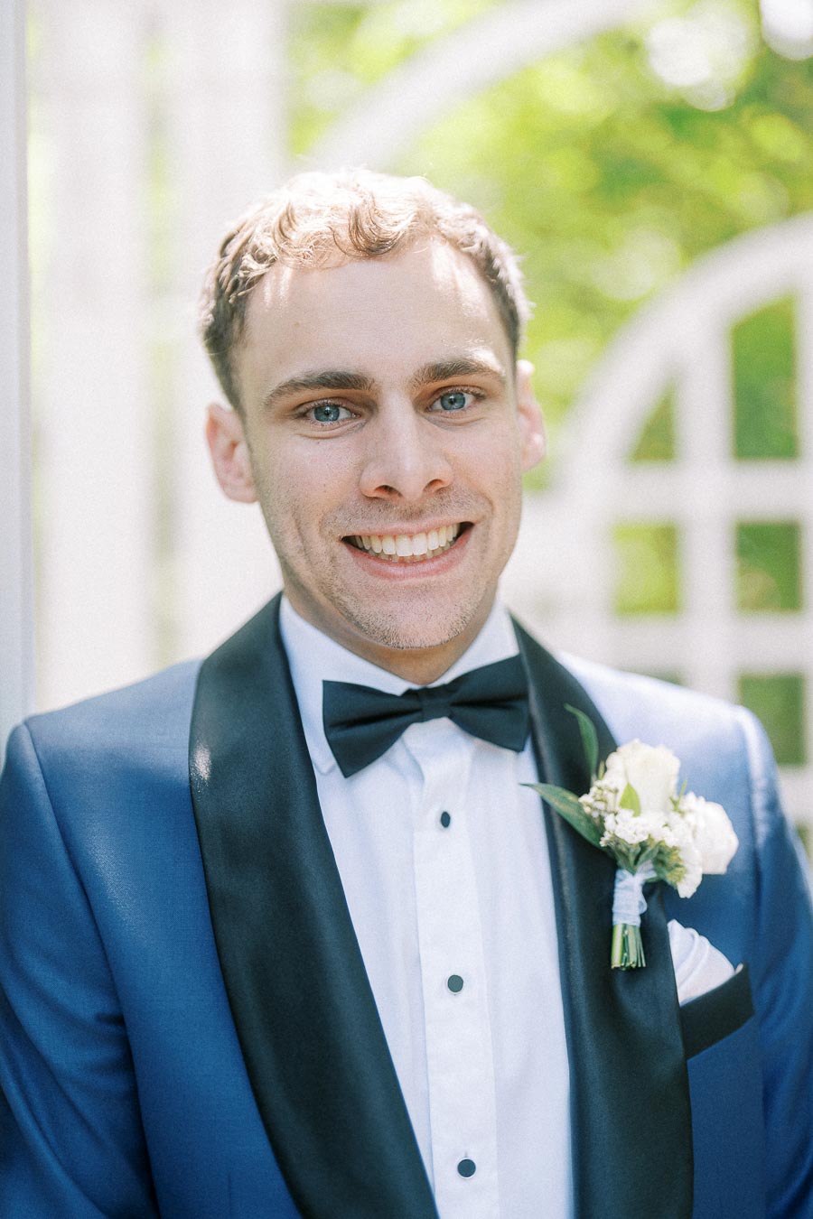 Rawle smiles in a blue tuxedo with black bow tie and white boutonniere outdoors at Ancaster Mill.