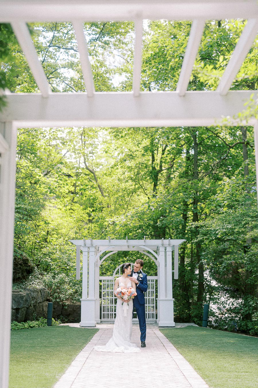 Bride and groom stand together under a white garden arbor surrounded by green trees at Ancaster Mill.