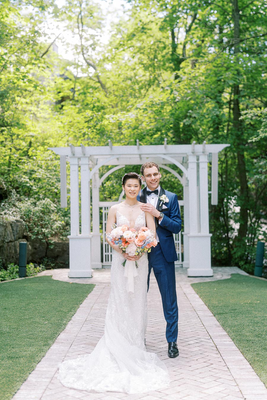 Miffy and Rawle stand together on a garden walkway before a white arbor at Ancaster Mill.