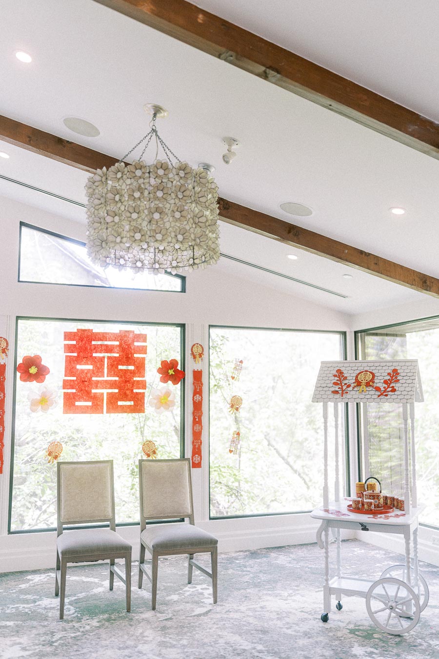 Bright tea ceremony room with two chairs, red double happiness decorations, and a dessert cart by large windows.