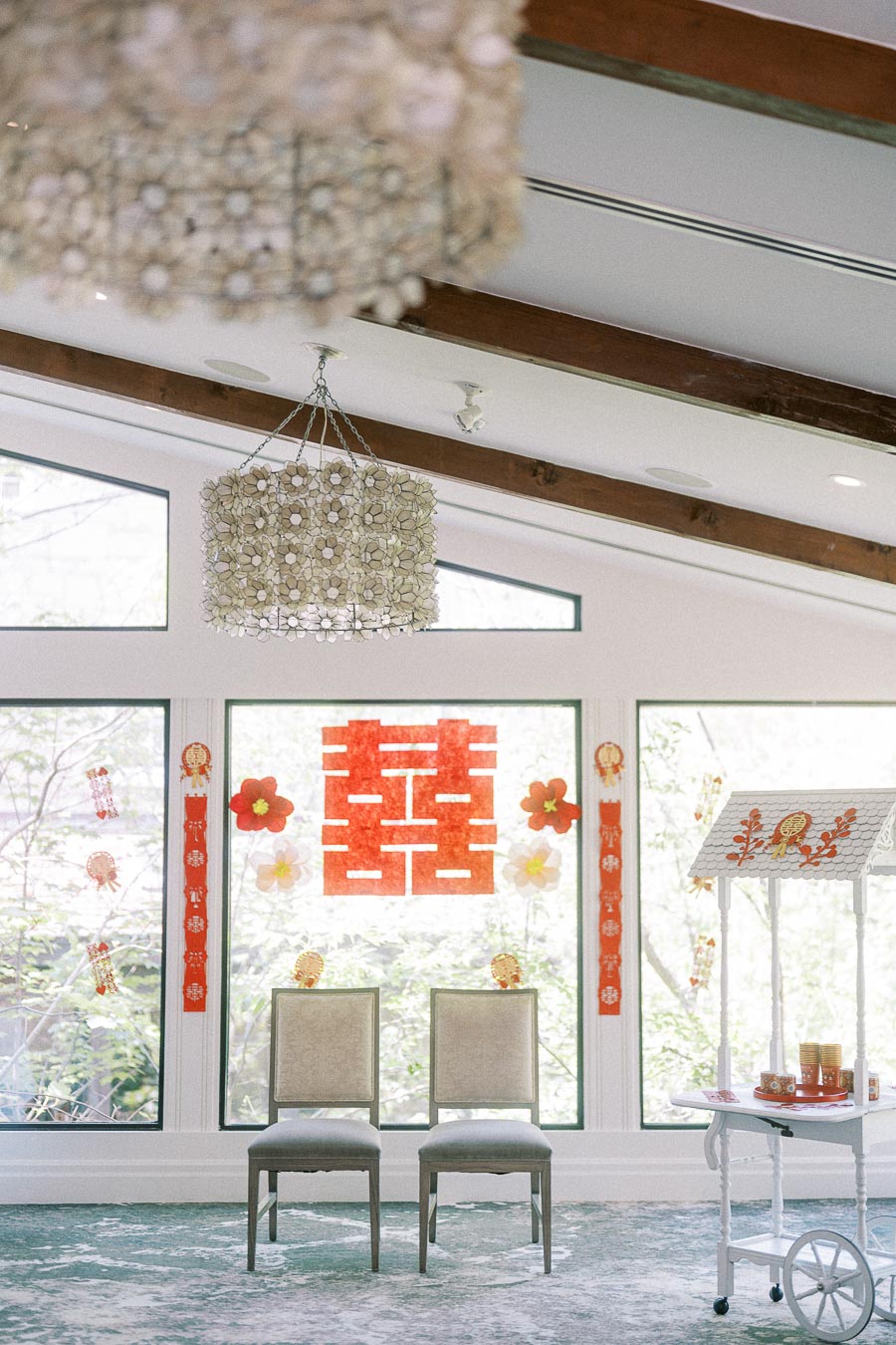 Chinese tea ceremony setup with double happiness decor, two chairs, tea cart and red ornaments by tall windows.