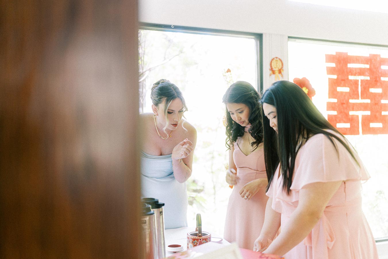Bride and bridesmaids in pink dresses gather near a window with red double happiness decorations