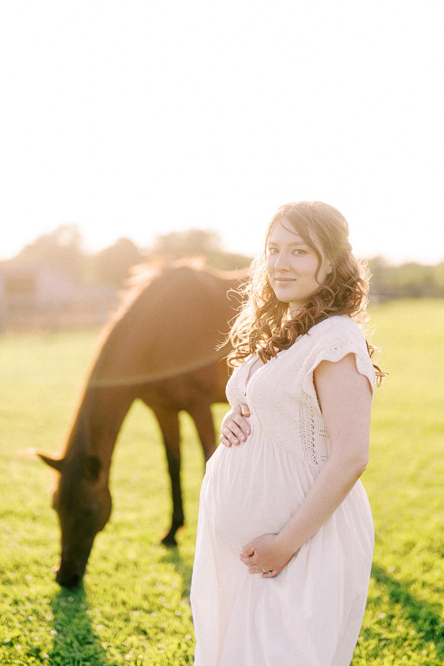 Pregnant woman in a white dress standing in a pasture with a grazing horse