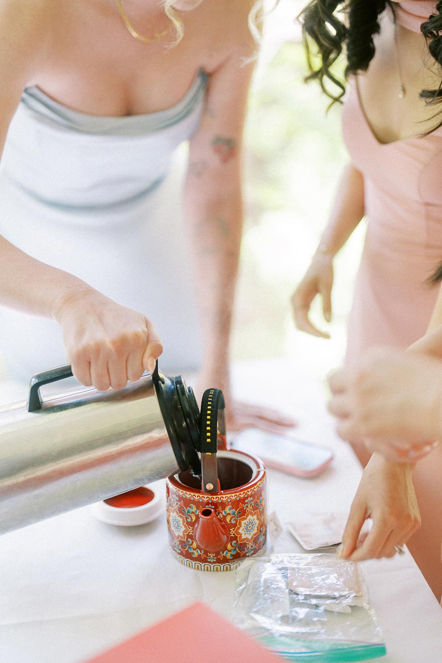 Women pour hot tea into a red patterned teapot during a Chinese tea ceremony setup.