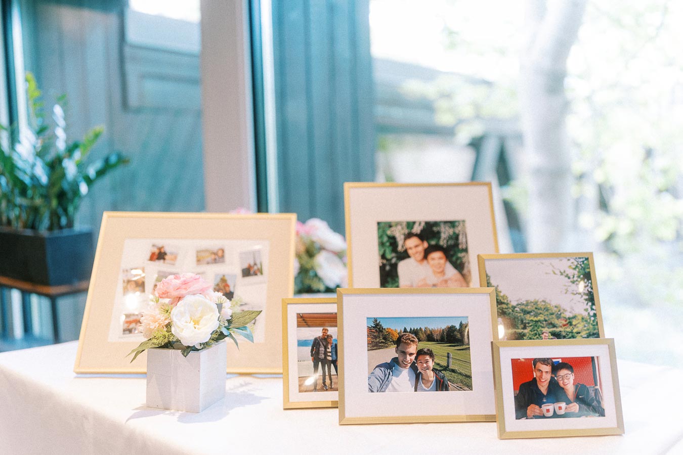 Framed couple photos and a small floral arrangement displayed on a white table by large windows.