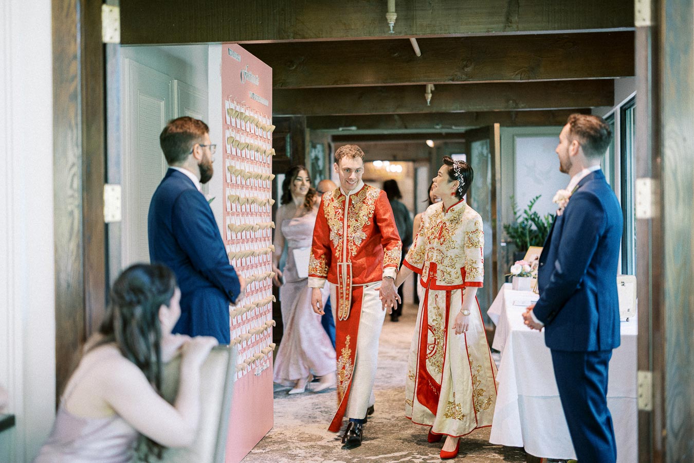 Miffy and Rawle walk hand in hand in red and gold Chinese wedding attire past a pink escort display.