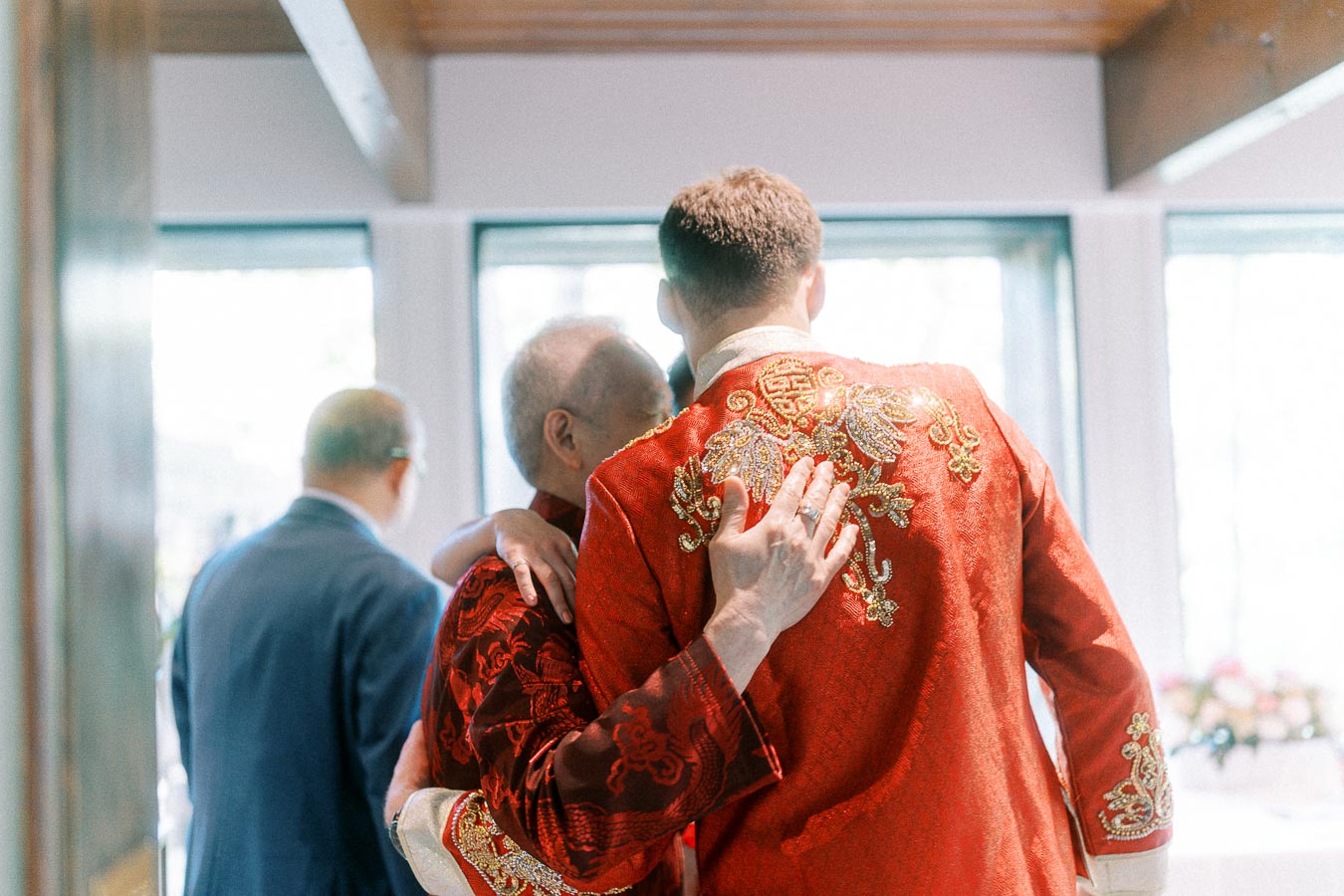 Groom in a red embroidered jacket embraces an elder during a Chinese tea ceremony indoors.