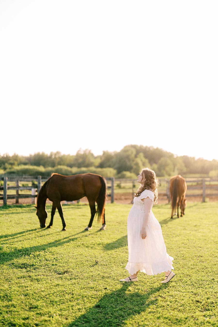 Pregnant woman in a white dress walking near grazing horses at The Green Acres