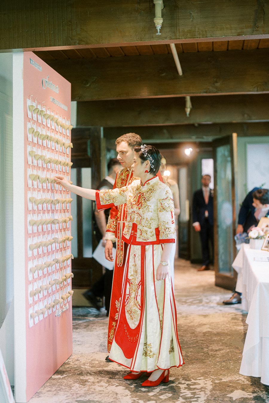 Miffy and Rawle in red and gold Chinese wedding attire pointing at a pink escort card display indoors