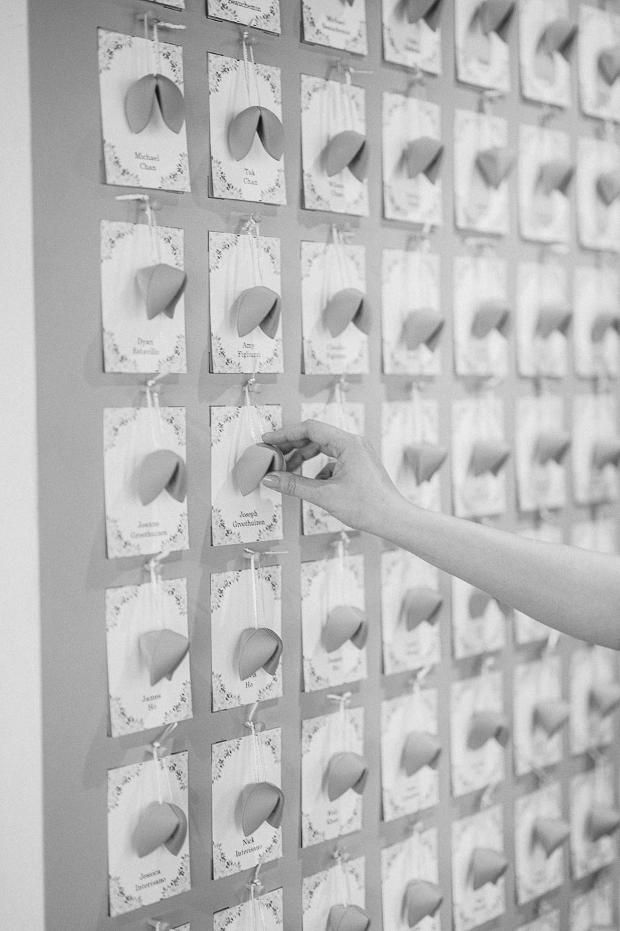 Hand selecting a fortune cookie escort card from a wall display of wedding guest place cards.