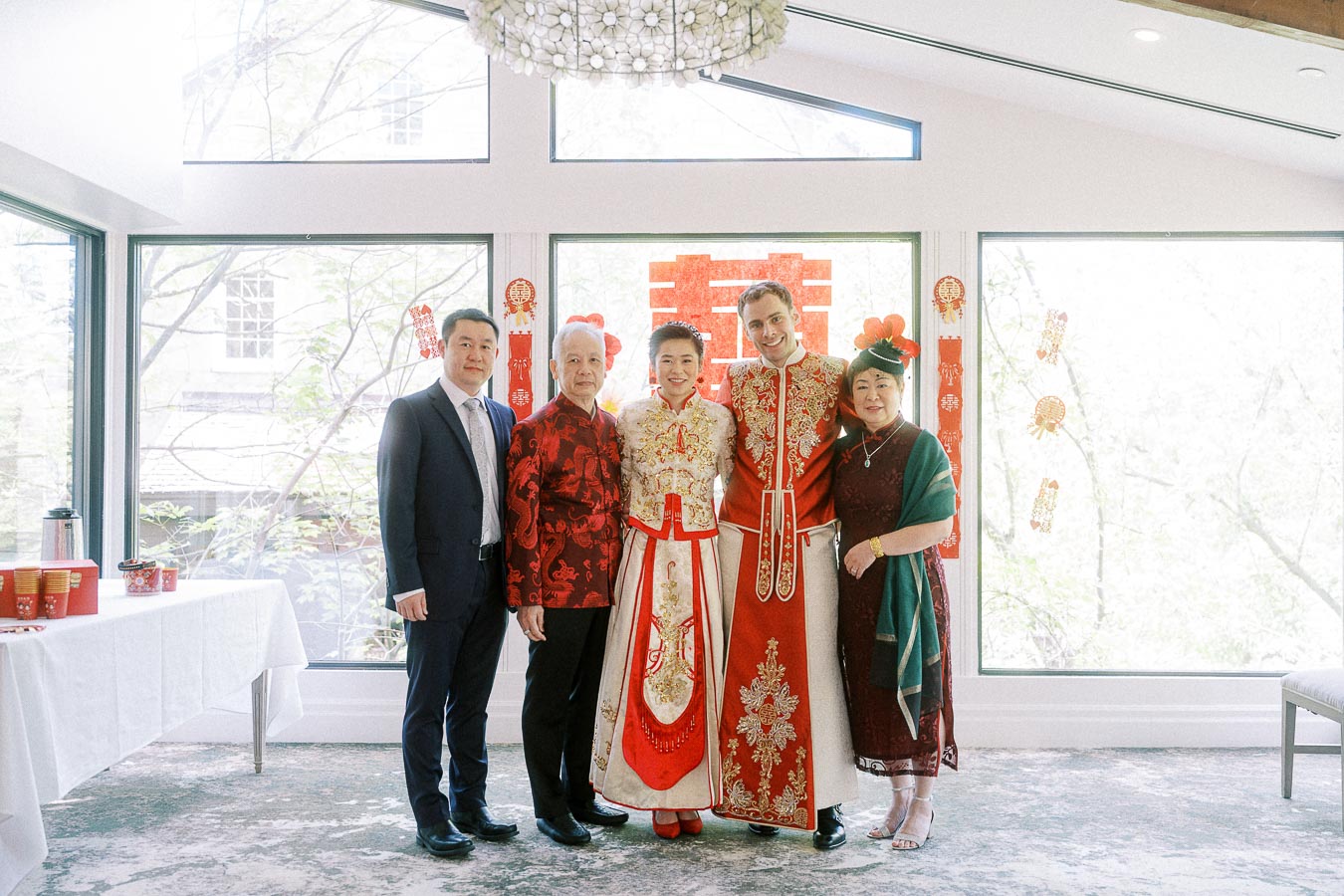 Miffy and Rawle pose with three family members in traditional red Chinese wedding attire by bright windows.