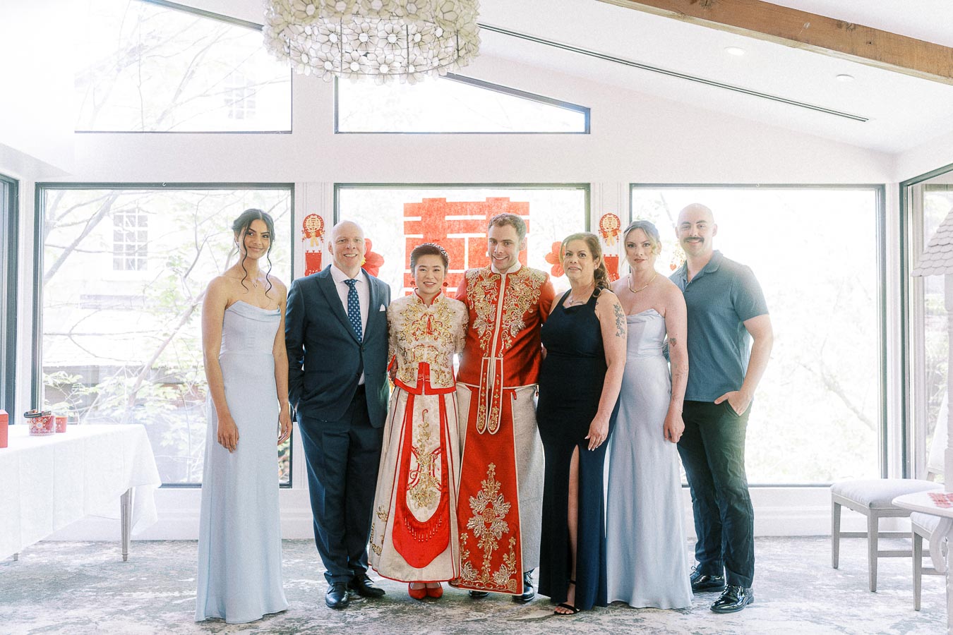 Miffy and Rawle in red Chinese wedding attire pose with family in a bright room with a double happiness sign.