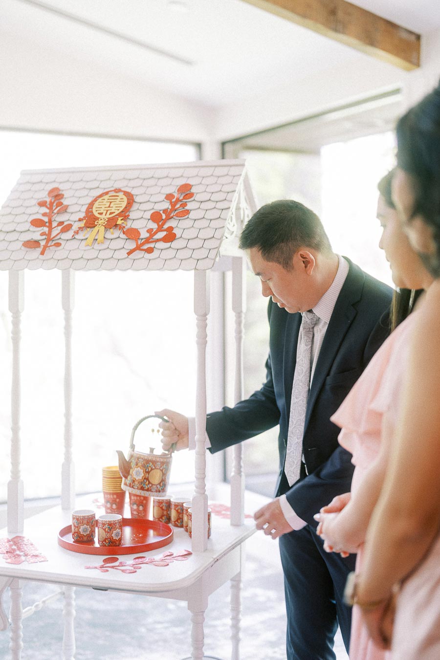 Man in a suit pours tea at a decorated Chinese tea ceremony station as guests watch nearby.