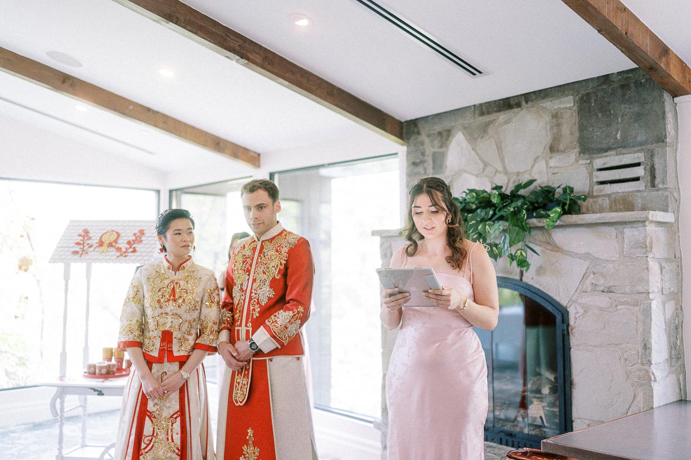 Miffy and Rawle in red Chinese wedding attire as a woman reads from a tablet beside a stone fireplace.