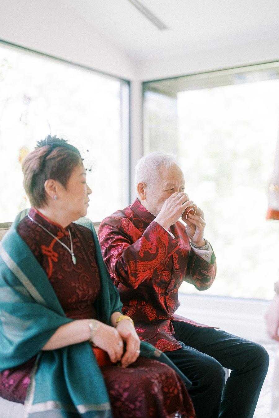 Elder in a red embroidered jacket drinks tea beside a woman in traditional attire near bright windows.