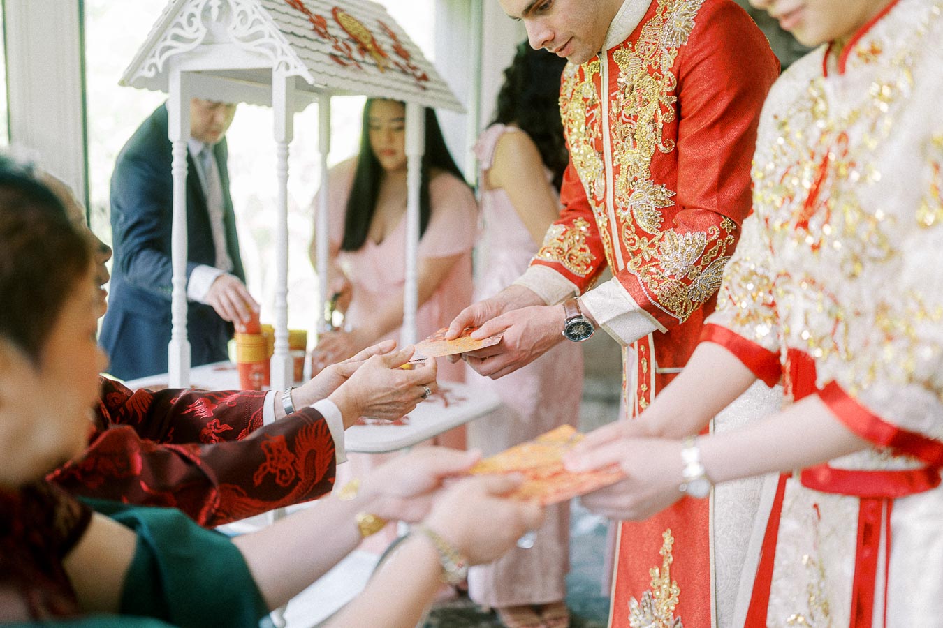 Couple in red and gold Chinese wedding attire offer red envelopes to seated family during a tea ceremony.