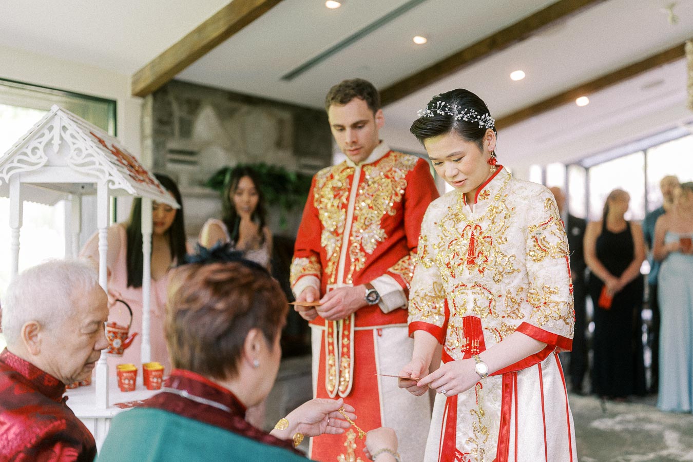 Miffy and Rawle in red and gold Chinese wedding attire serve tea to seated elders during their tea ceremony.