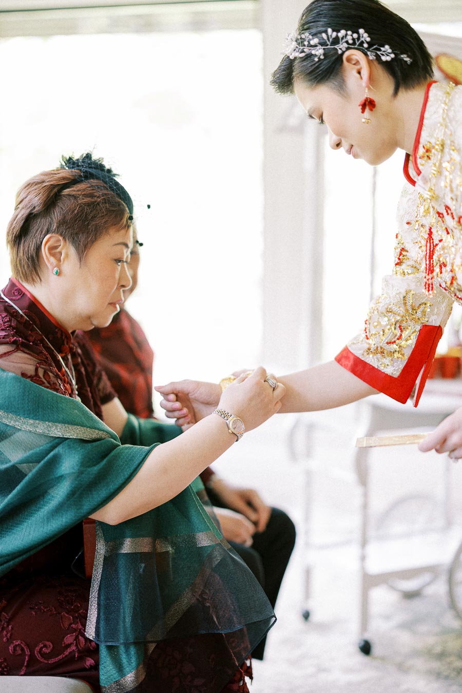 Bride in a red and gold Chinese dress offers tea to a seated woman during a tea ceremony.