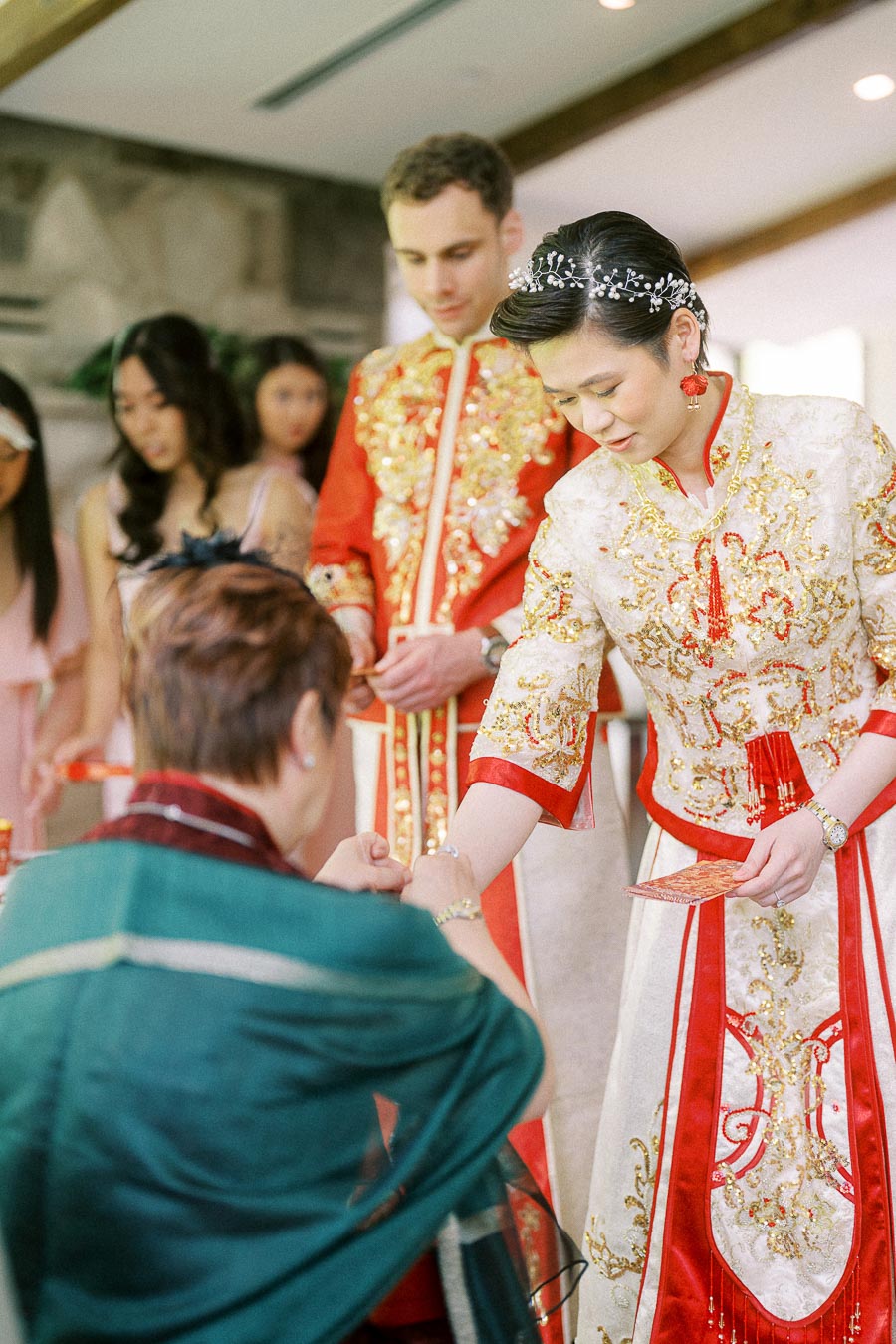 Bride in embroidered Chinese wedding attire greets an elder during a tea ceremony as the groom stands behind her.