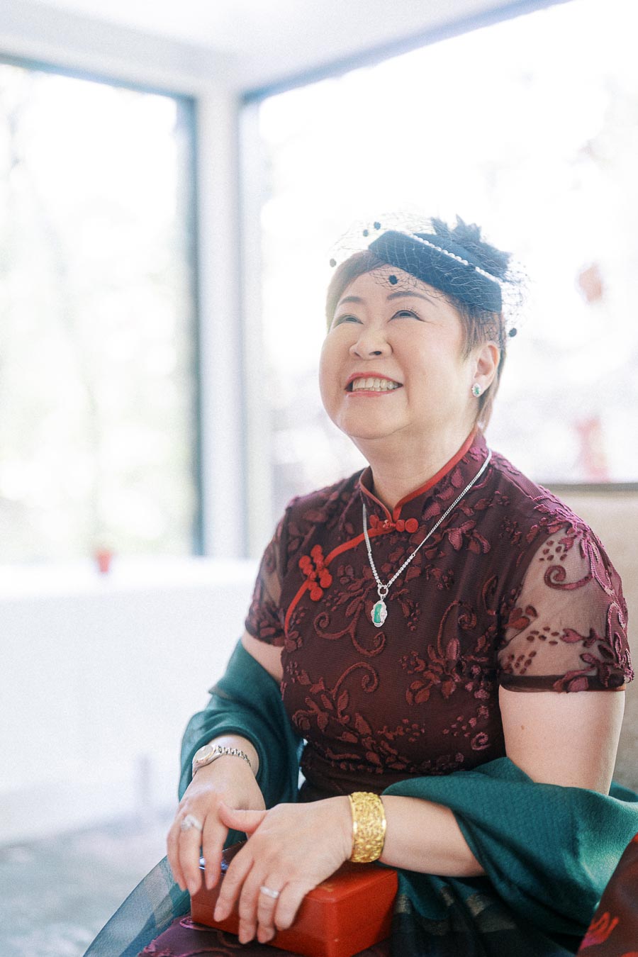Smiling woman in a burgundy qipao and black fascinator seated by bright windows at a wedding.