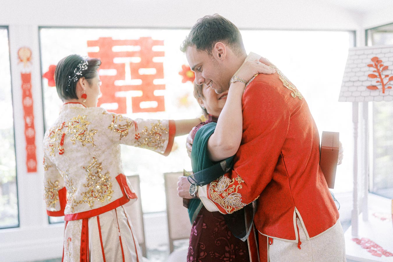 Bride in embroidered Chinese attire watches groom in red jacket hug an elder during a tea ceremony