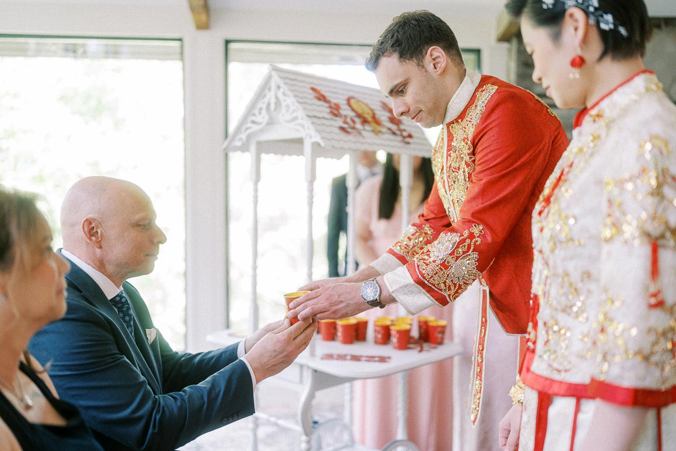 Rawle in a red embroidered jacket offers tea to a seated elder during a Chinese tea ceremony.