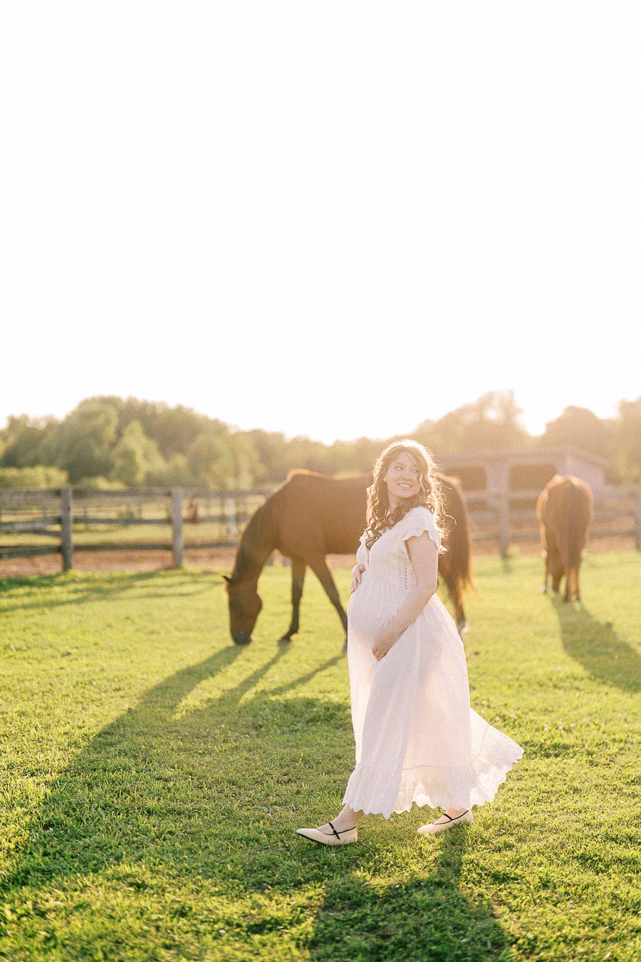Pregnant woman in a white dress standing in a pasture with grazing horses
