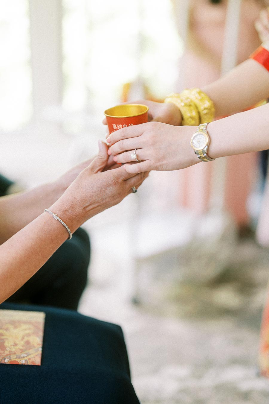 Hands passing a red tea cup during a Chinese tea ceremony, with gold bracelets and wedding rings visible.