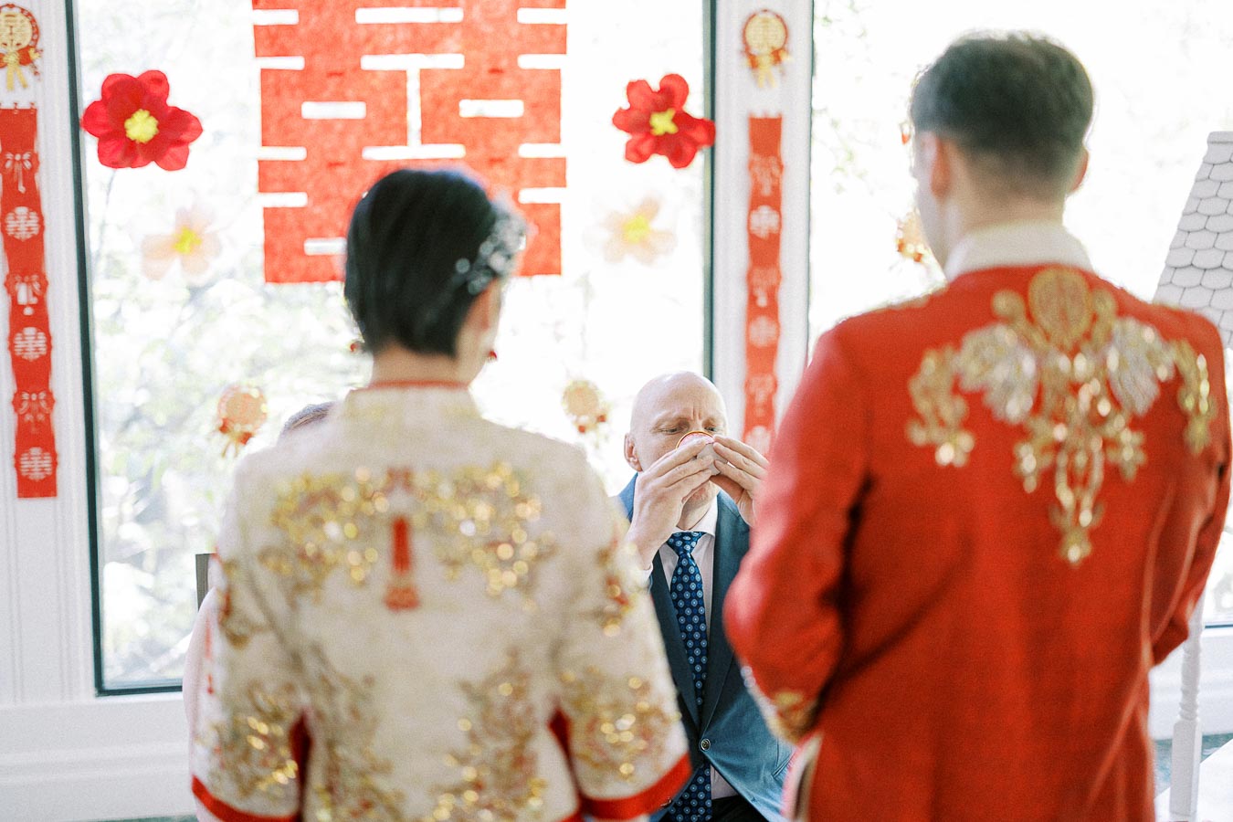 Bride and groom in embroidered Chinese wedding attire face a seated man drinking tea during a tea ceremony.