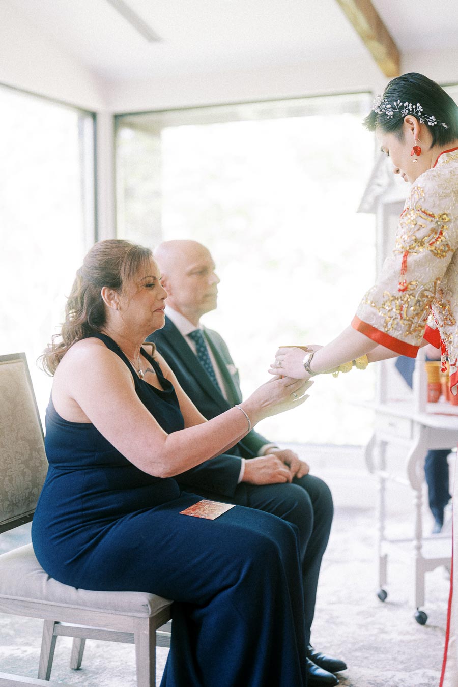 Bride in red and gold Chinese wedding attire serves tea to seated elders during a tea ceremony.
