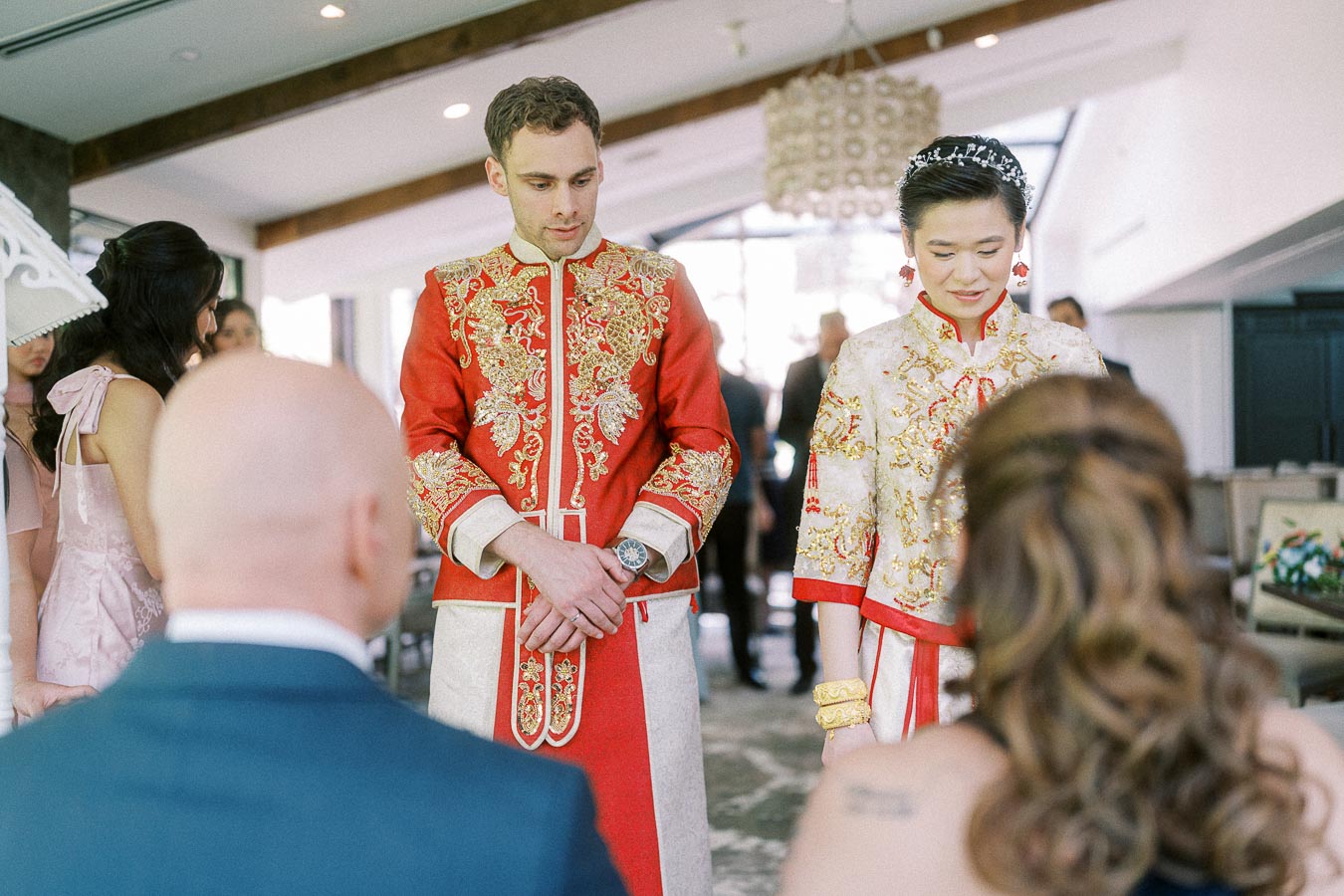 Miffy and Rawle stand in red and gold Chinese wedding attire before seated elders during a tea ceremony.