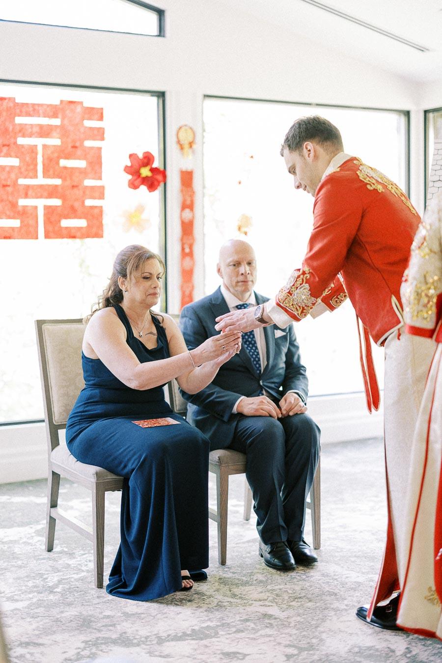 Groom in red Chinese wedding attire serves tea to seated parents beside a red double happiness window sign.