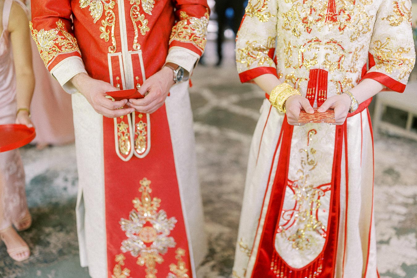 Couple in red and gold Chinese wedding outfits holding red envelopes during a tea ceremony