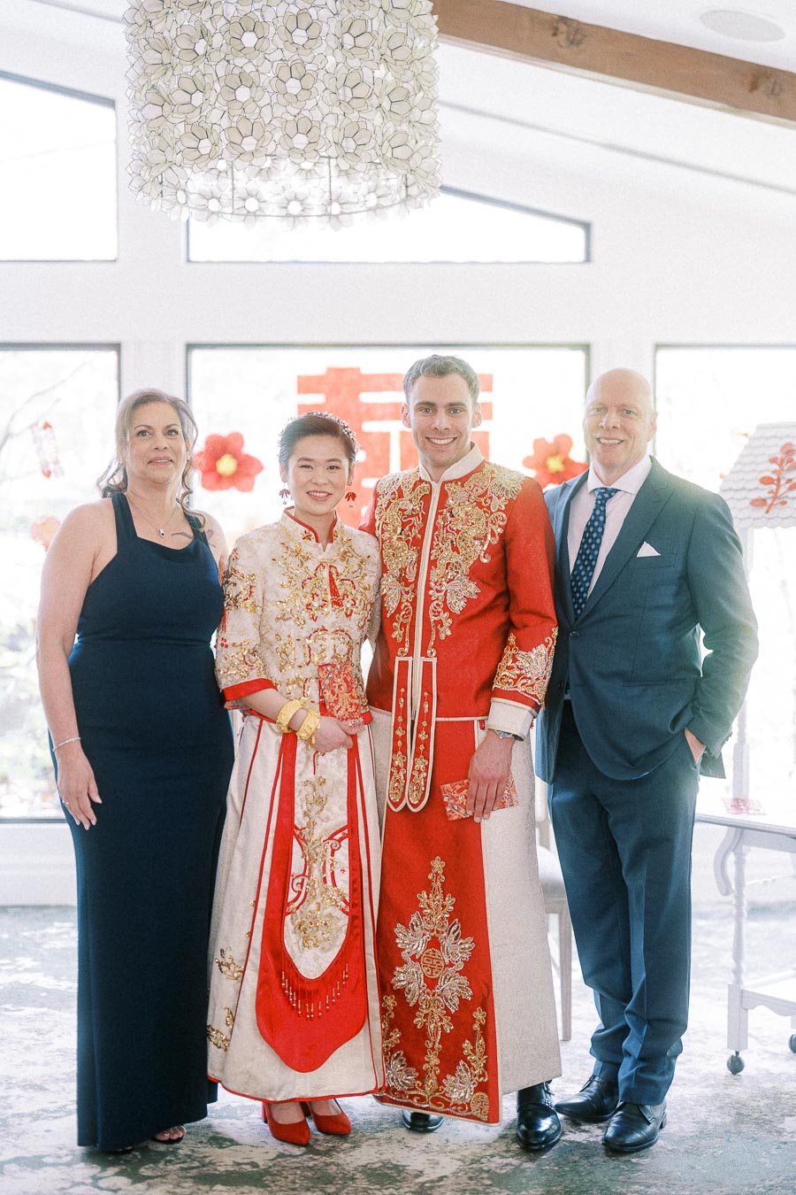Miffy and Rawle in red and gold Chinese wedding attire standing with two family members indoors.