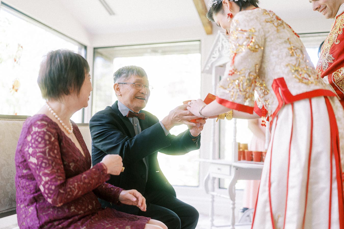 Bride and groom in traditional Chinese wedding attire present a red envelope to smiling elders during tea ceremony.