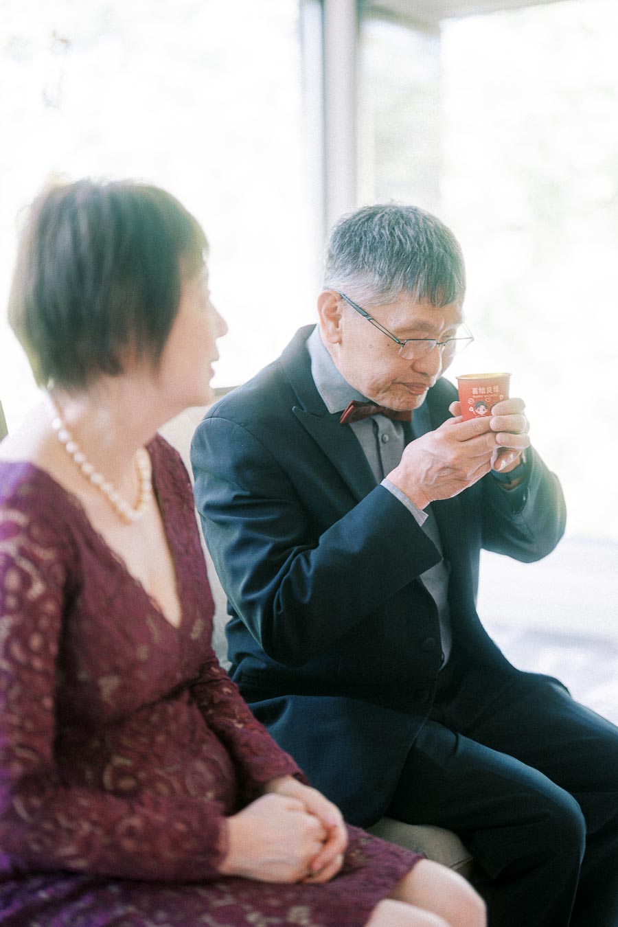 Older man in a suit drinks tea from a red cup while seated beside a woman in a burgundy dress.