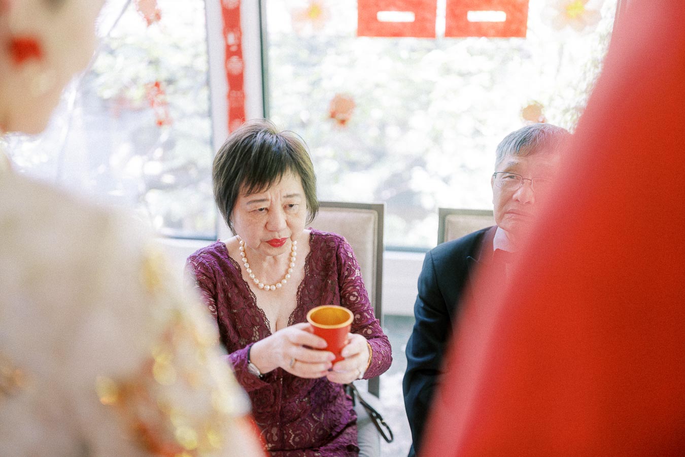 Woman in a burgundy dress holds a red tea cup during a Chinese tea ceremony beside a seated man.