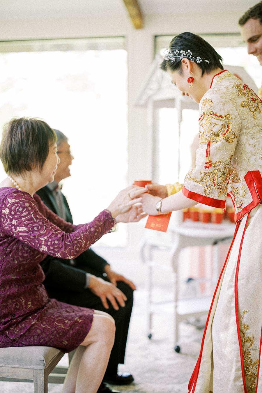 Bride in embroidered red and gold Chinese dress offers tea and a red envelope to seated elders during tea ceremony.