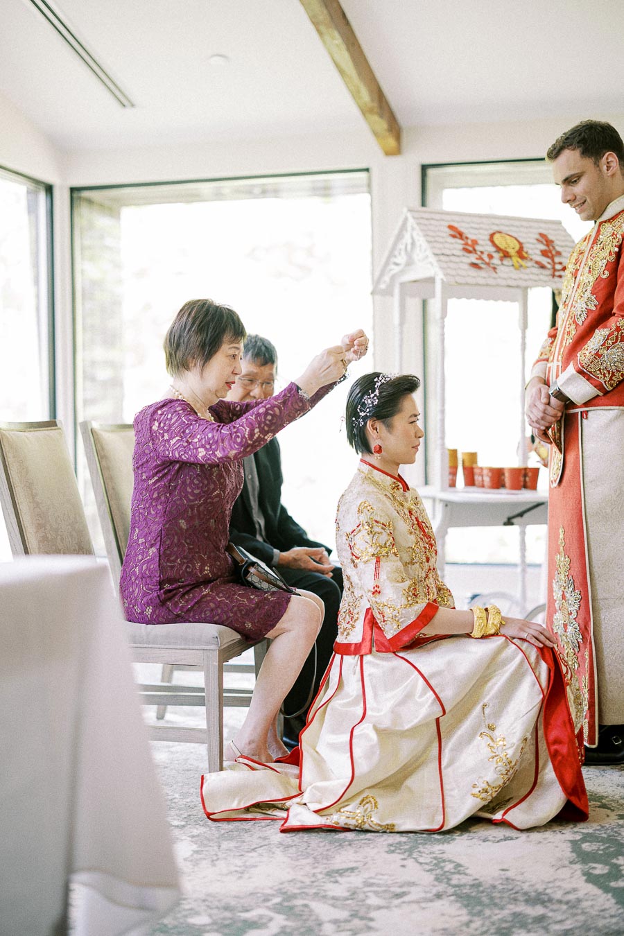 Bride kneels in a white and red Chinese wedding dress while an elder places jewelry in her hair.