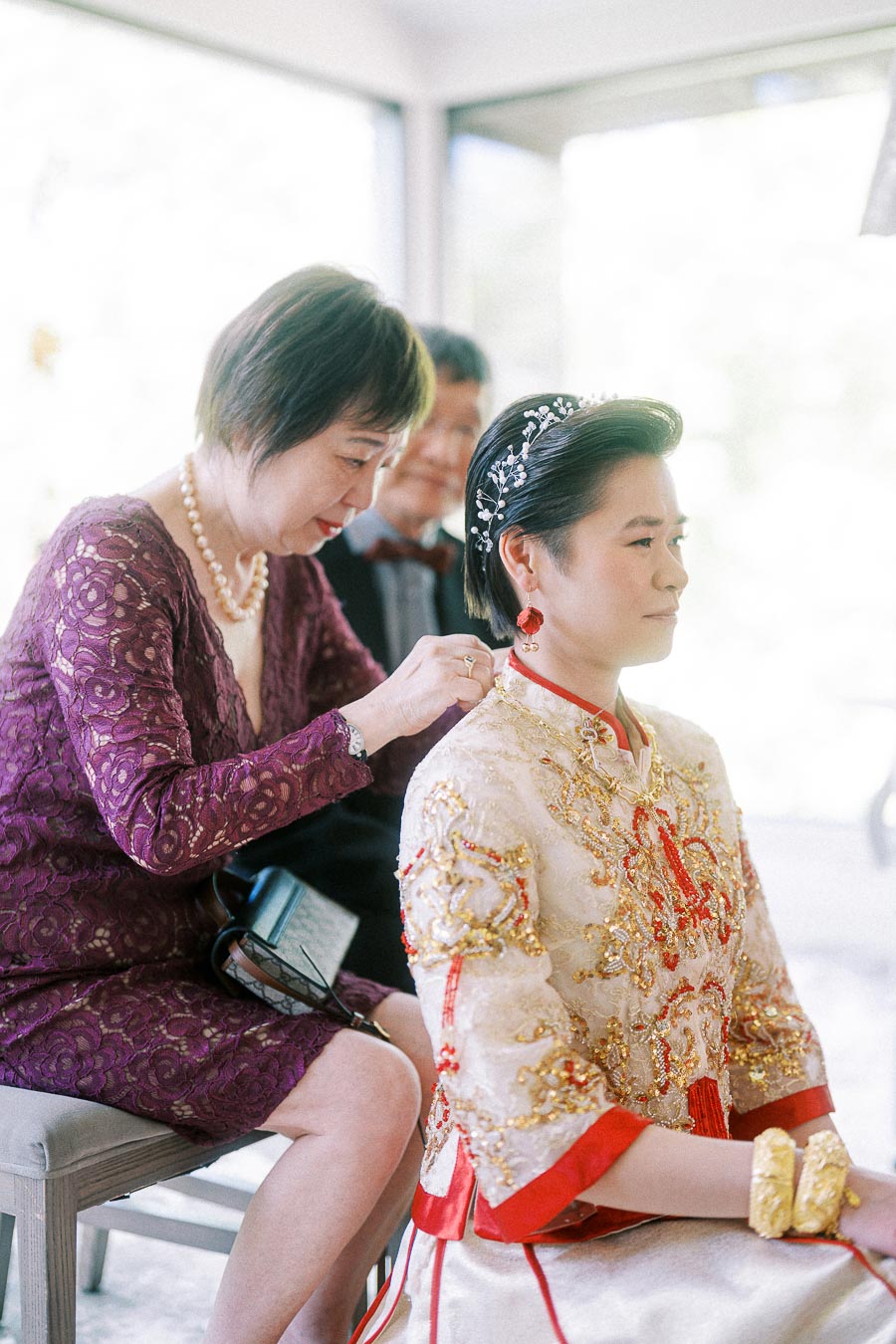 Woman fastening Miffy's ornate Chinese wedding jacket as she sits in a bright room with hair jewelry